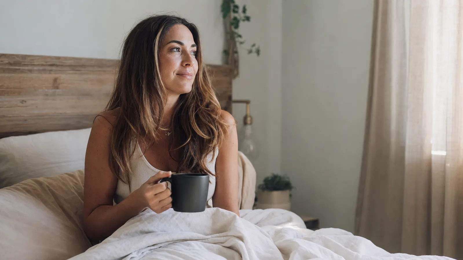 Woman sitting up in bed in the morning looking rested, holding a mug of coffee, sunlight through curtains